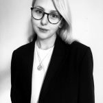 A young woman with blonde hair and glasses. She wears a smart casual suit and looks forward.