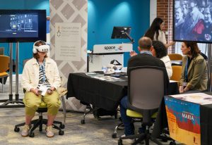 A person wearing a VR headset sits in front of a display screen at a conference booth, while others engage in discussion at a nearby table.