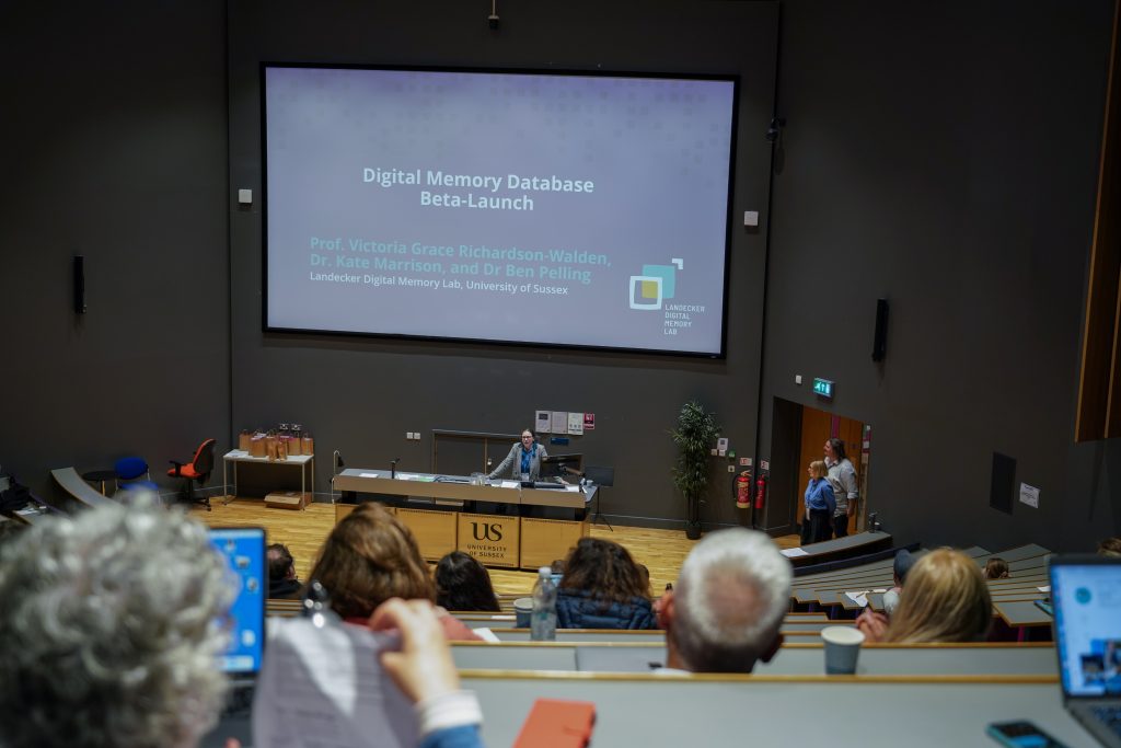 A presenter speaks in a lecture hall during the Digital Memory Database Beta-Launch, with a projection screen displaying names of project leads from the Landecker Digital Memory Lab at the University of Sussex.