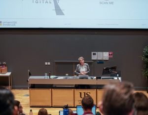 A speaker stands at a lecture hall podium presenting to an audience, with a large screen displaying a QR code and graphics related to digital Holocaust research.