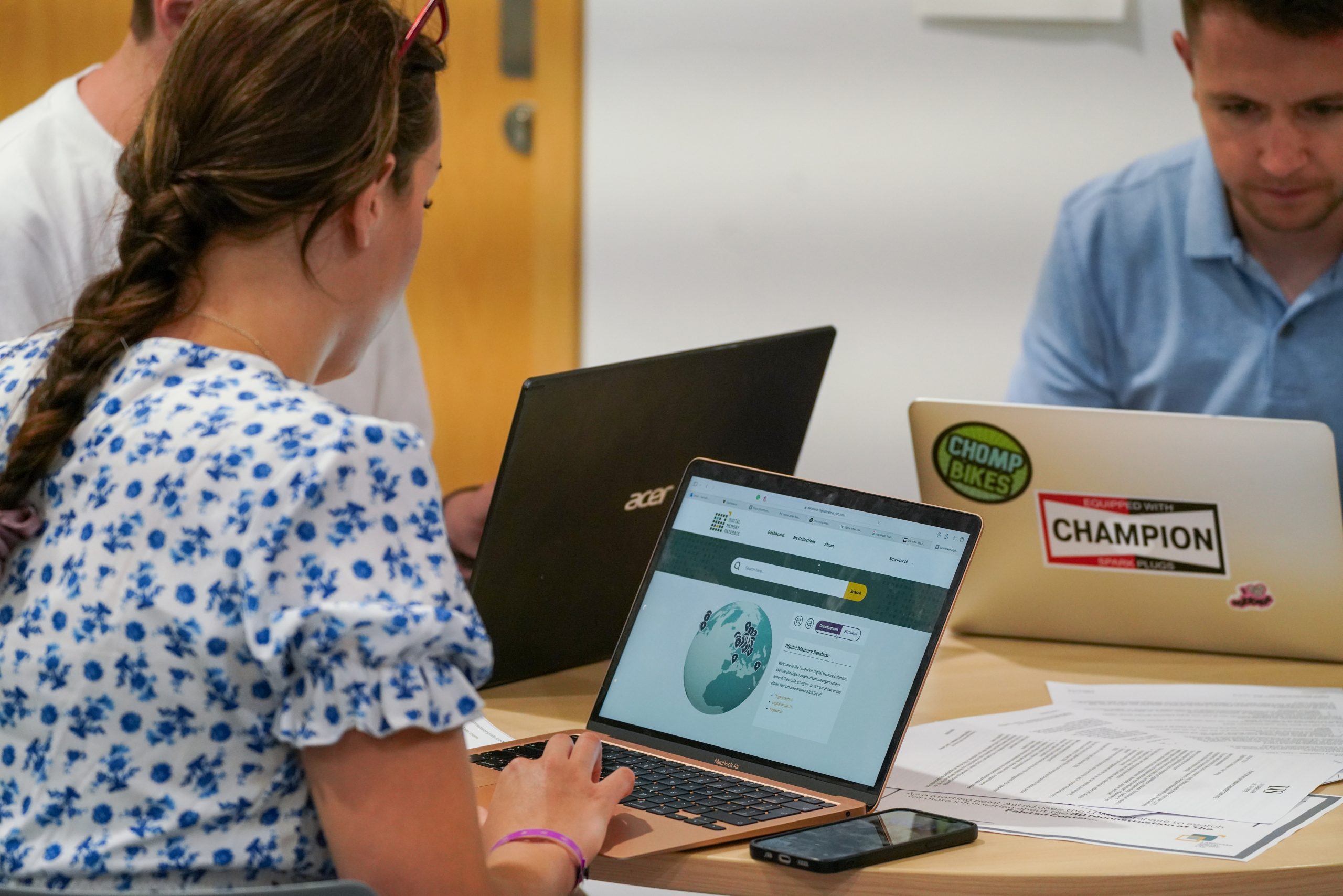 People working at a table with laptops, one of which displays the Digital Memory Database interface on the screen.