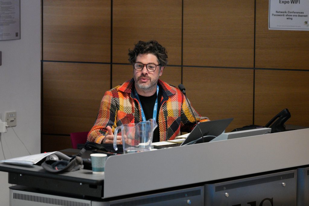 A man wearing glasses and a colorful checkered jacket speaks at a podium with a microphone and laptop during a conference.