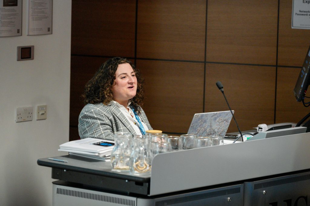 A woman with curly hair and a plaid blazer speaks at a podium with a microphone and laptop during a conference session.