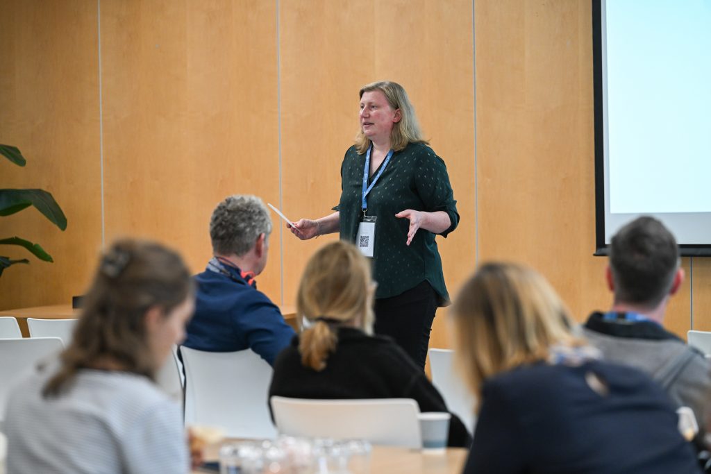 A woman holding a paper speaks to an audience seated at tables during a presentation in a conference room.