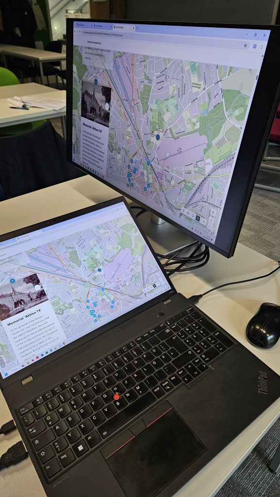 Laptop connected to a monitor displaying a map interface with marked points, suggesting an interactive Holocaust memory or historical mapping project.