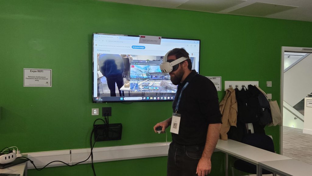 A man wearing a VR headset interacts with a digital display on a green wall screen during a demonstration, possibly exploring a virtual Holocaust memory experience.