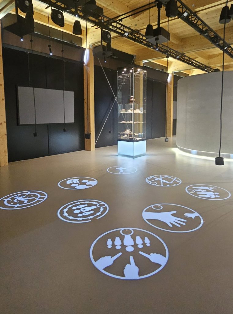 Modern exhibition space with wooden ceiling beams, interactive floor projections displaying symbolic icons, and a central glass display case containing historical artifacts, illuminated by overhead lights.