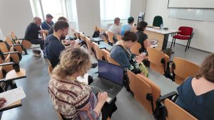 Participants seated in a classroom or lecture hall, using laptops, tablets, and smartphones during a workshop or seminar session.