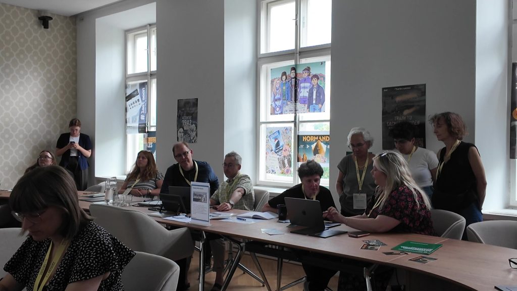 Group of people seated and standing around tables in a workshop room, engaging with laptops and printed materials, with educational posters on the windows and walls.